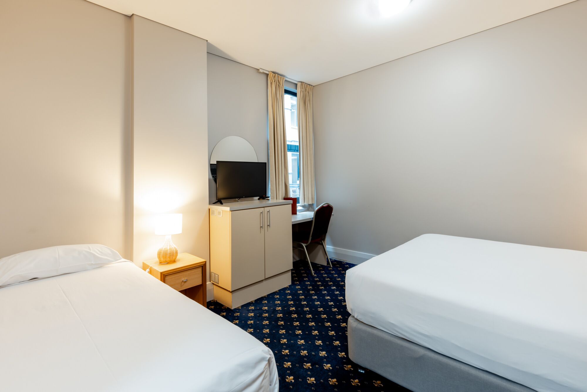 Habitación de hotel con dos camas individuales, un escritorio con silla, televisor y un mueble bajo.
