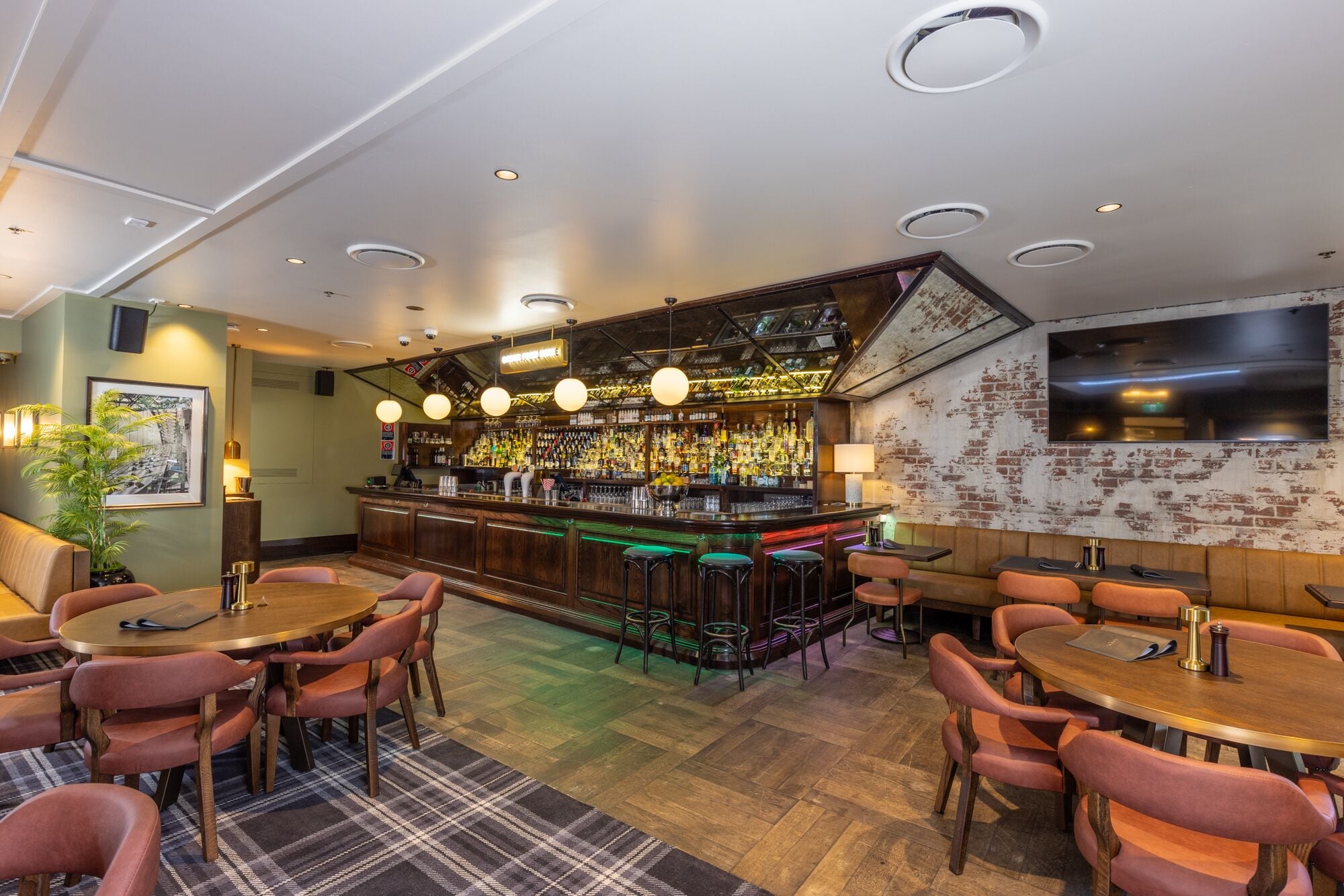 A bar area with a wooden bar counter, multiple stools, and a variety of bottles. There are tables with chairs for seating.