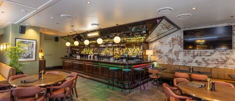 A bar area with a wooden bar counter, multiple stools, and a variety of bottles. There are tables with chairs for seating.