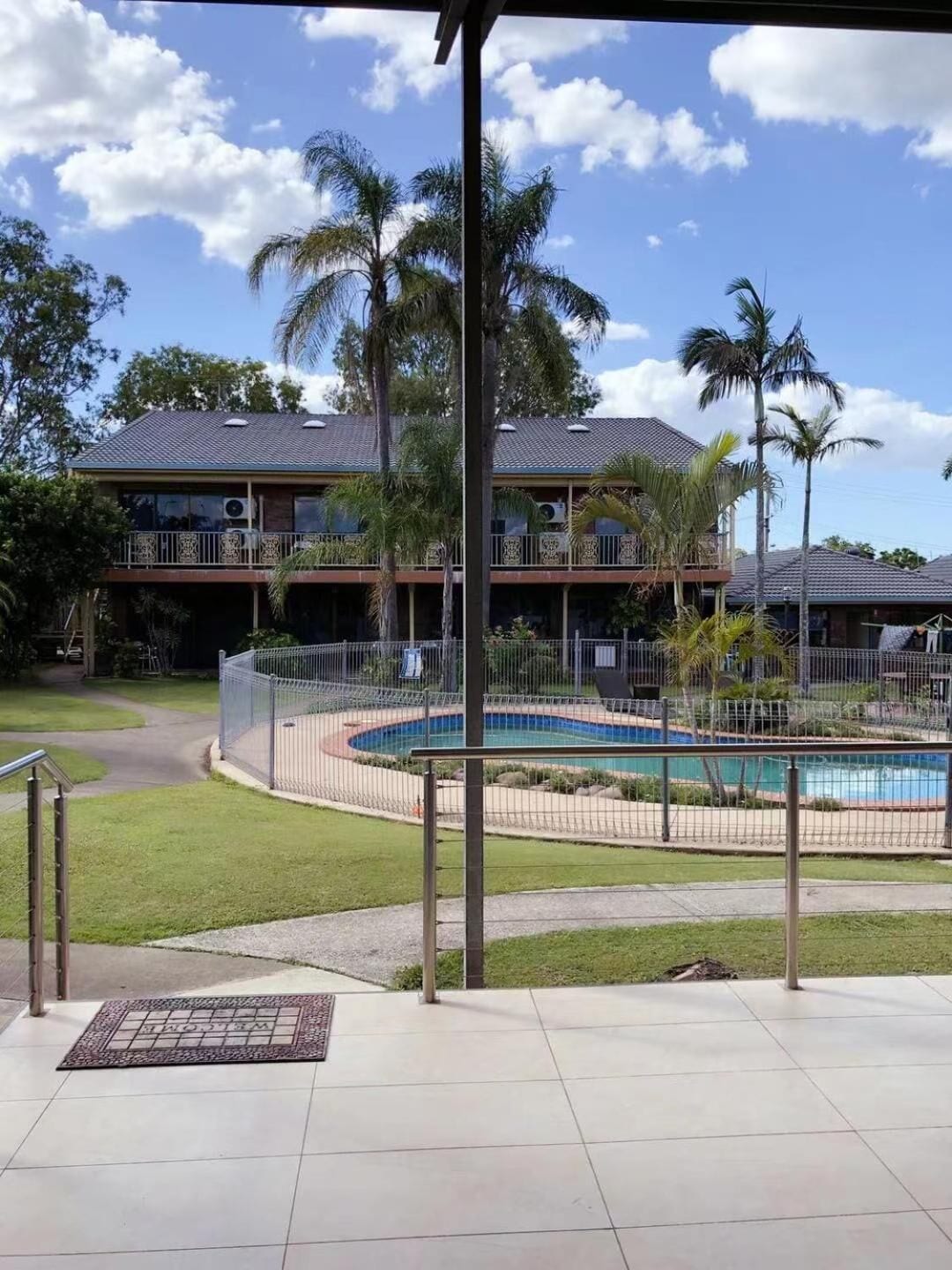A two-story building with a swimming pool, surrounded by a fence and palm trees.