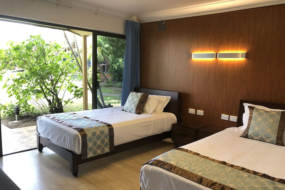 A hotel room with two beds, a large window with a view of greenery, and a wooden wall with built-in lighting.