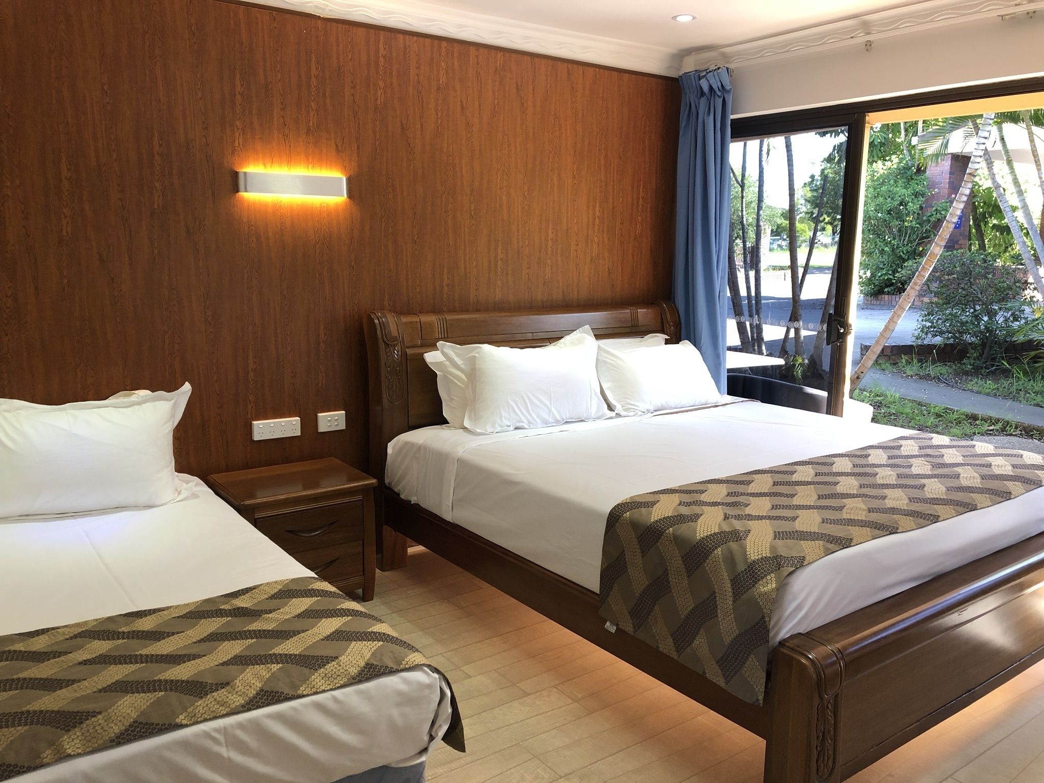 Een hotelkamer met twee bedden, houten paneelwanden, een nachtkastje, een wandlamp en uitzicht op de omgeving.