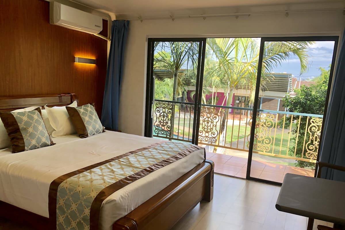 A hotel room with a large bed, a wooden headboard, a balcony with a view of palm trees and a building, and a table and chair in the corner.