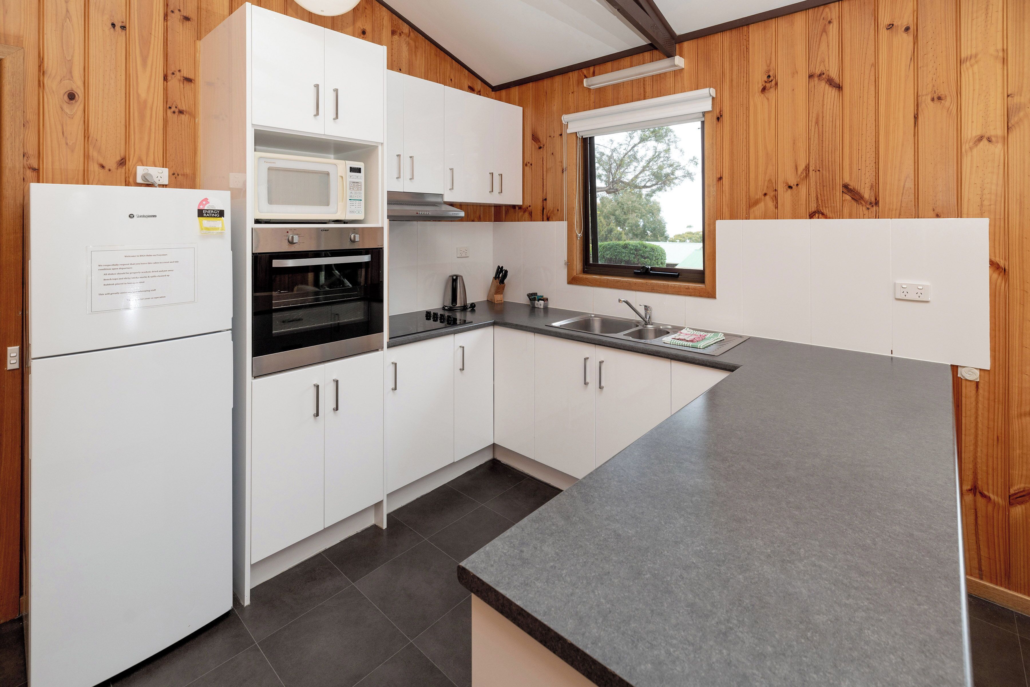 A kitchen with white cabinets, a microwave, an oven, a sink, and a window with a view.