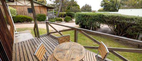A wooden deck with a round table and four chairs, overlooking a well-maintained garden and a building with a green roof.