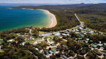 An aerial view of a coastal area with a beach, dense forest, and a cluster of buildings with green roofs.