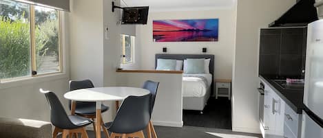 A compact, modern room with a dining area, a bed with a headboard, a kitchen with a black range hood, and a large window with a view of greenery.