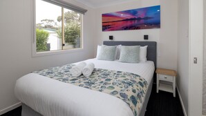 Una cama bien hecha con colcha floral y dos almohadas, una mesita de noche con cajón, una lámpara de noche y un ventanal con vistas a árboles y una casa.