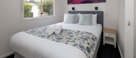 Una cama bien hecha con colcha floral y dos almohadas, una mesita de noche con cajón, una lámpara de noche y un ventanal con vistas a árboles y una casa.