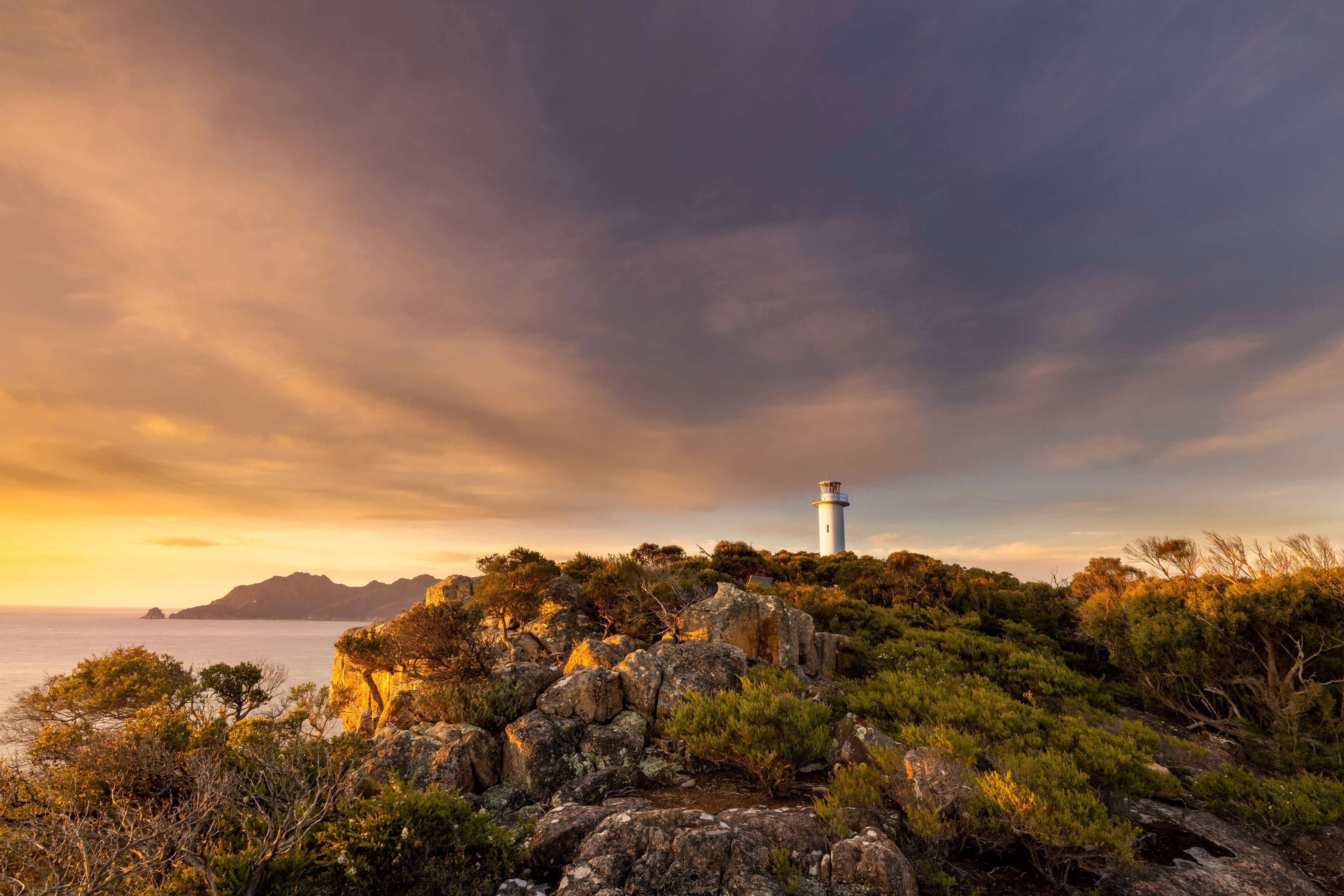 A lighthouse on a rocky outcrop with a view of the sea and distant mountains.