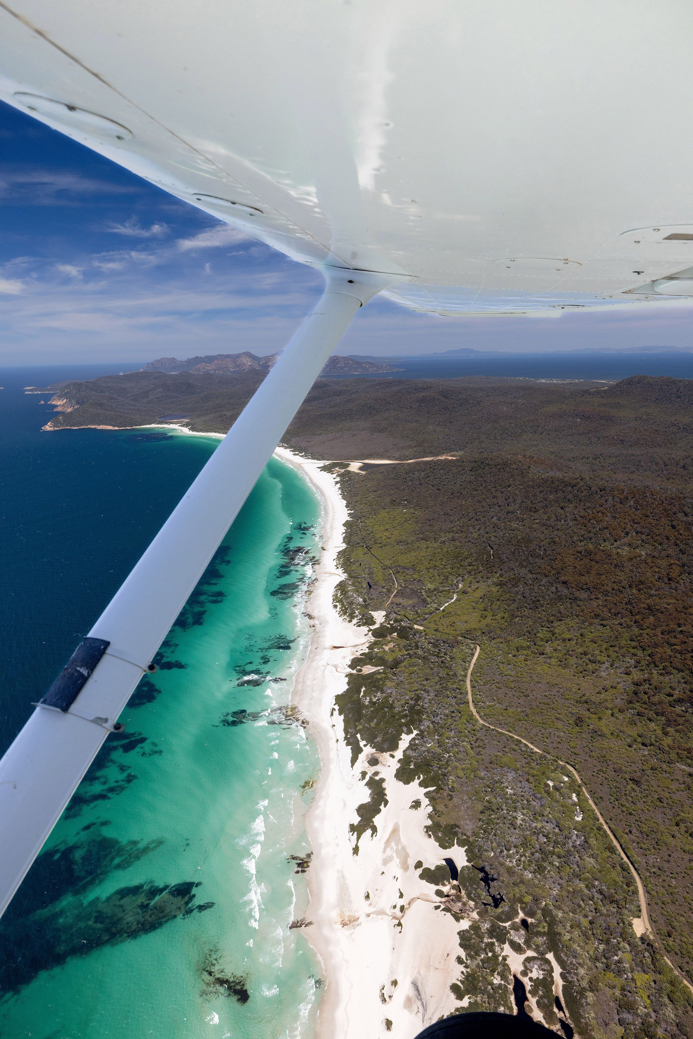An aerial view of a beach with clear turquoise waters, a sandy shoreline, and a forested area.