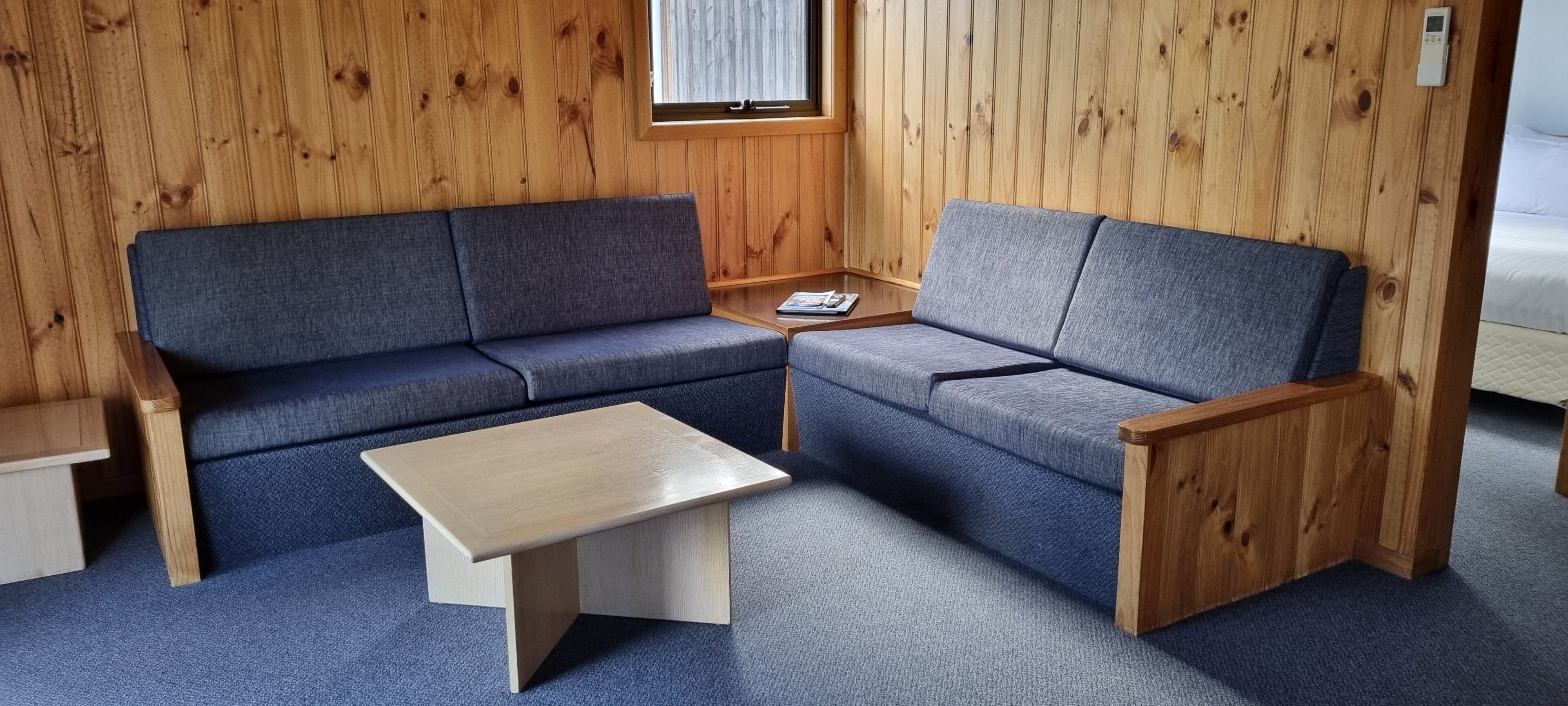 A room with a wooden paneled wall, a grey sofa, a wooden coffee table, and a small bedside table.