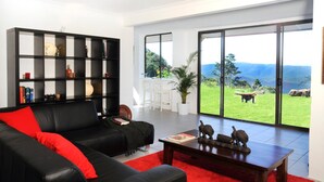 A modern living room with a black leather sofa, a red throw pillow, a wooden coffee table, and a black bookshelf. There is a large window with a view of a green landscape and a stone structure. - Clouds on Beechmont (Beechmont)