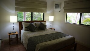 Ein ordentlich eingerichtetes Schlafzimmer mit einem Bett, zwei Nachttischlampen, einem Nachttisch und einem Fenster mit Blick auf Grünflächen.