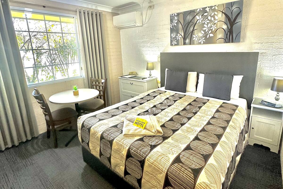 Habitación de hotel con cama, escritorio, silla, ventana con cortinas, unidad de aire acondicionado y ventilador de techo.