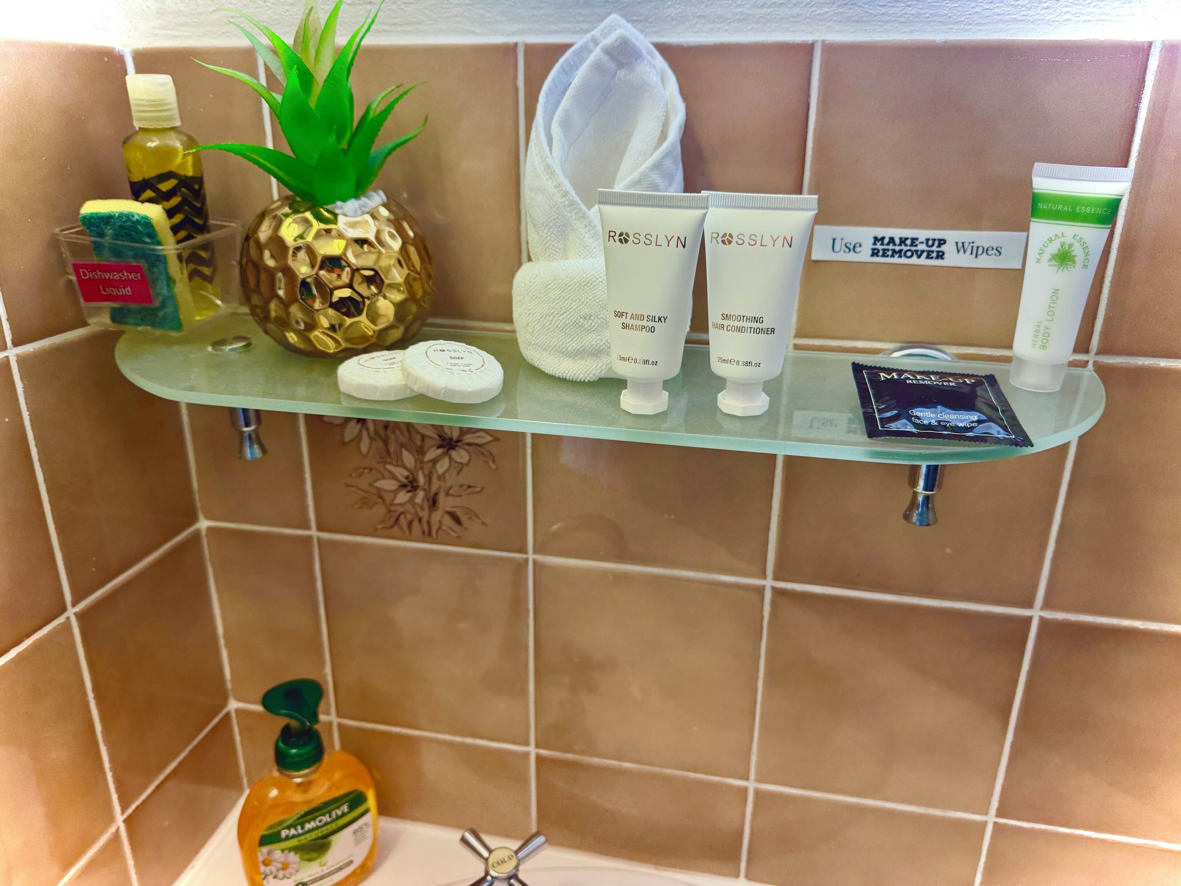 Un estante de baño con diversos artículos de aseo, incluyendo una botella de detergente, un dispensador de jabón con forma de piña, dos tubos de productos capilares Rosslyn, un paquete de toallitas desmaquillantes y una botella de aceite de oliva de palma.