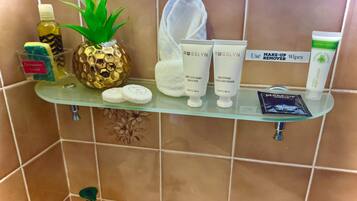 Un estante de baño con diversos artículos de aseo, incluyendo una botella de detergente, un dispensador de jabón con forma de piña, dos tubos de productos capilares Rosslyn, un paquete de toallitas desmaquillantes y una botella de aceite de oliva de palma.