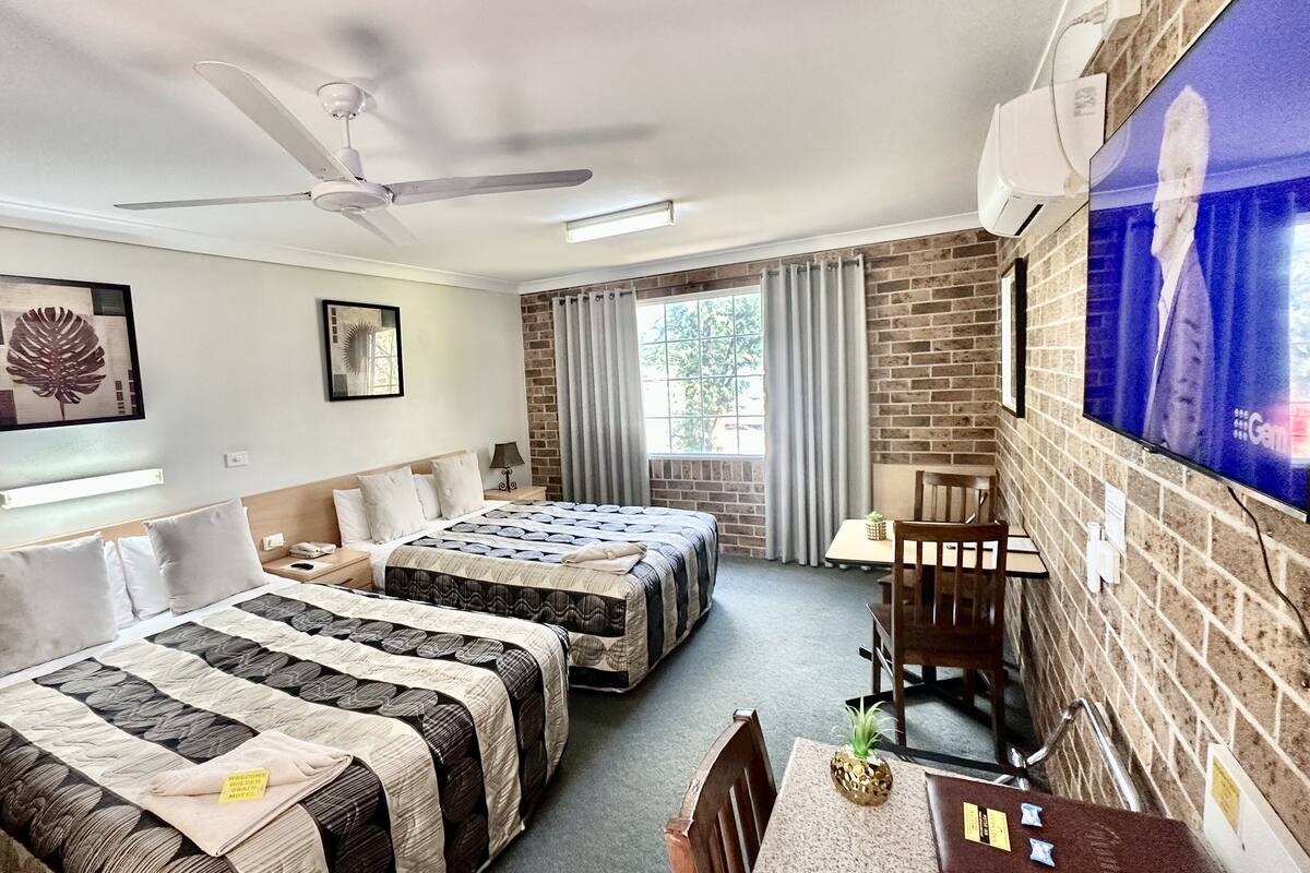 Habitación de hotel con dos camas, ventilador de techo, televisión y pared de ladrillo.