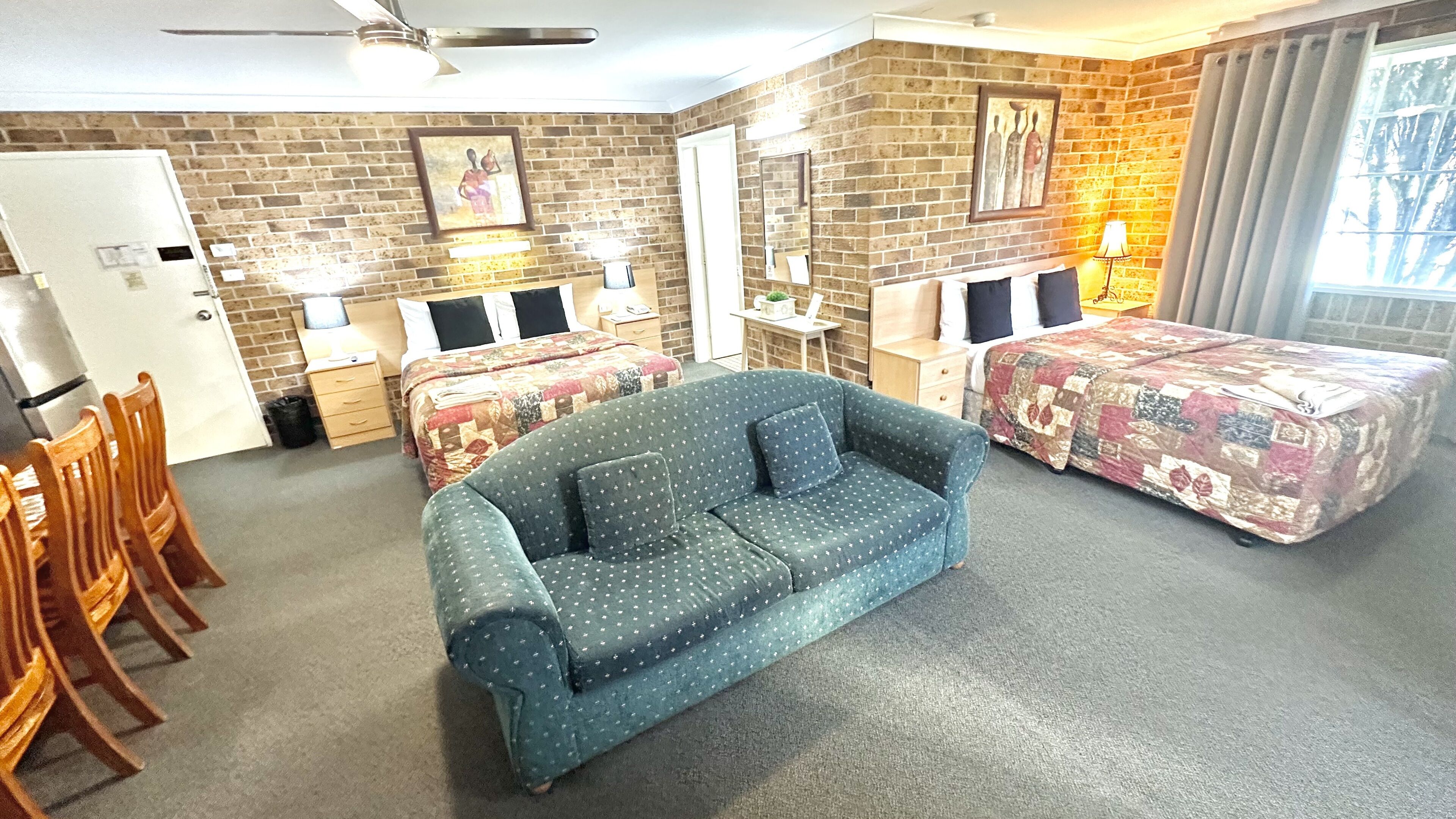 A room with a sofa, two beds with patterned bedding, a desk with a chair, a small table, and a refrigerator.