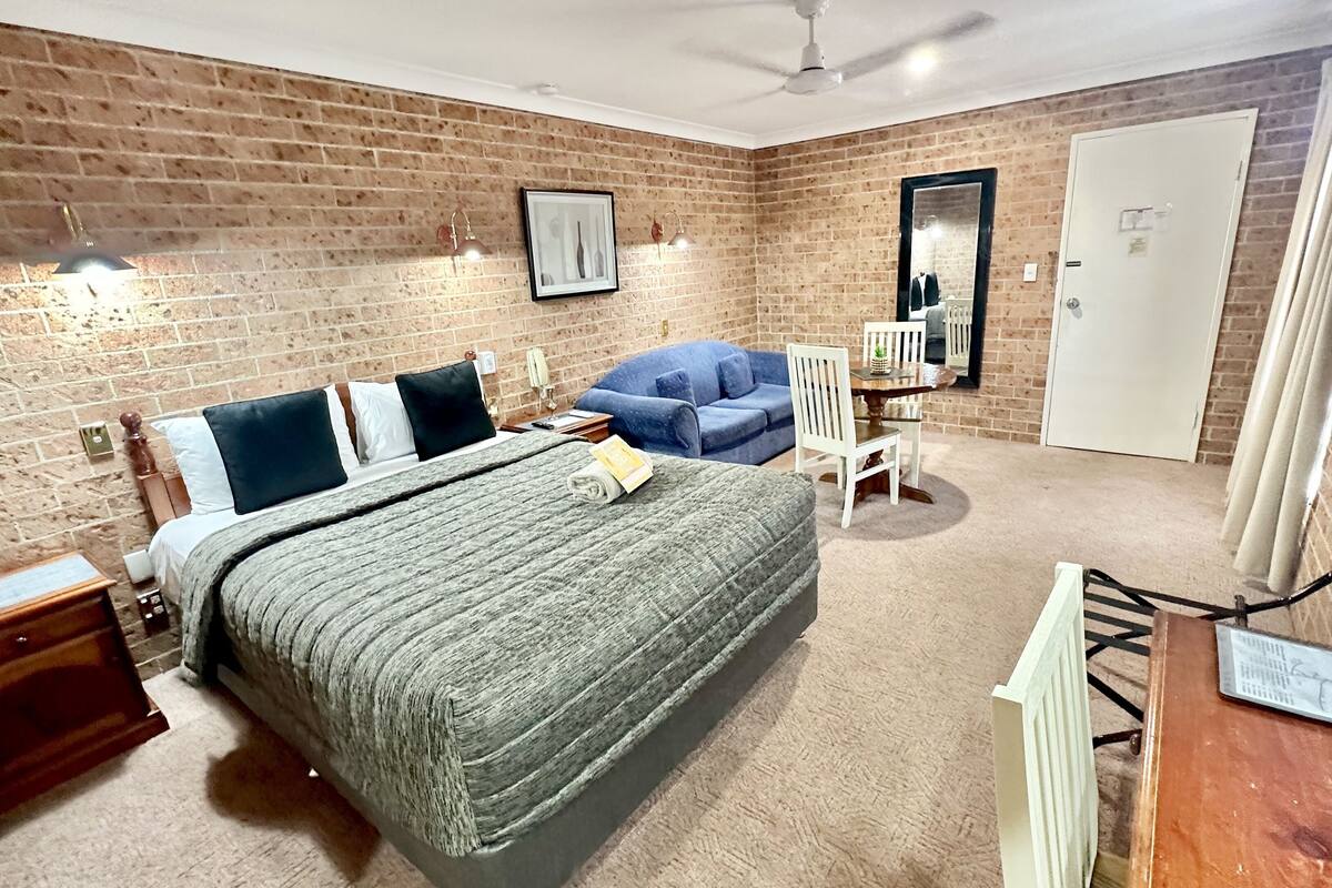 Un dormitorio con pared de ladrillo, una cama con manta gris, un sofá azul, una silla blanca, una mesita de noche de madera y una puerta blanca.