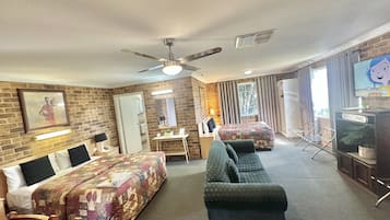 A hotel room with a brick wall, a ceiling fan, a bed with a floral bedspread, a sofa, a desk with a lamp, and a TV on a stand.