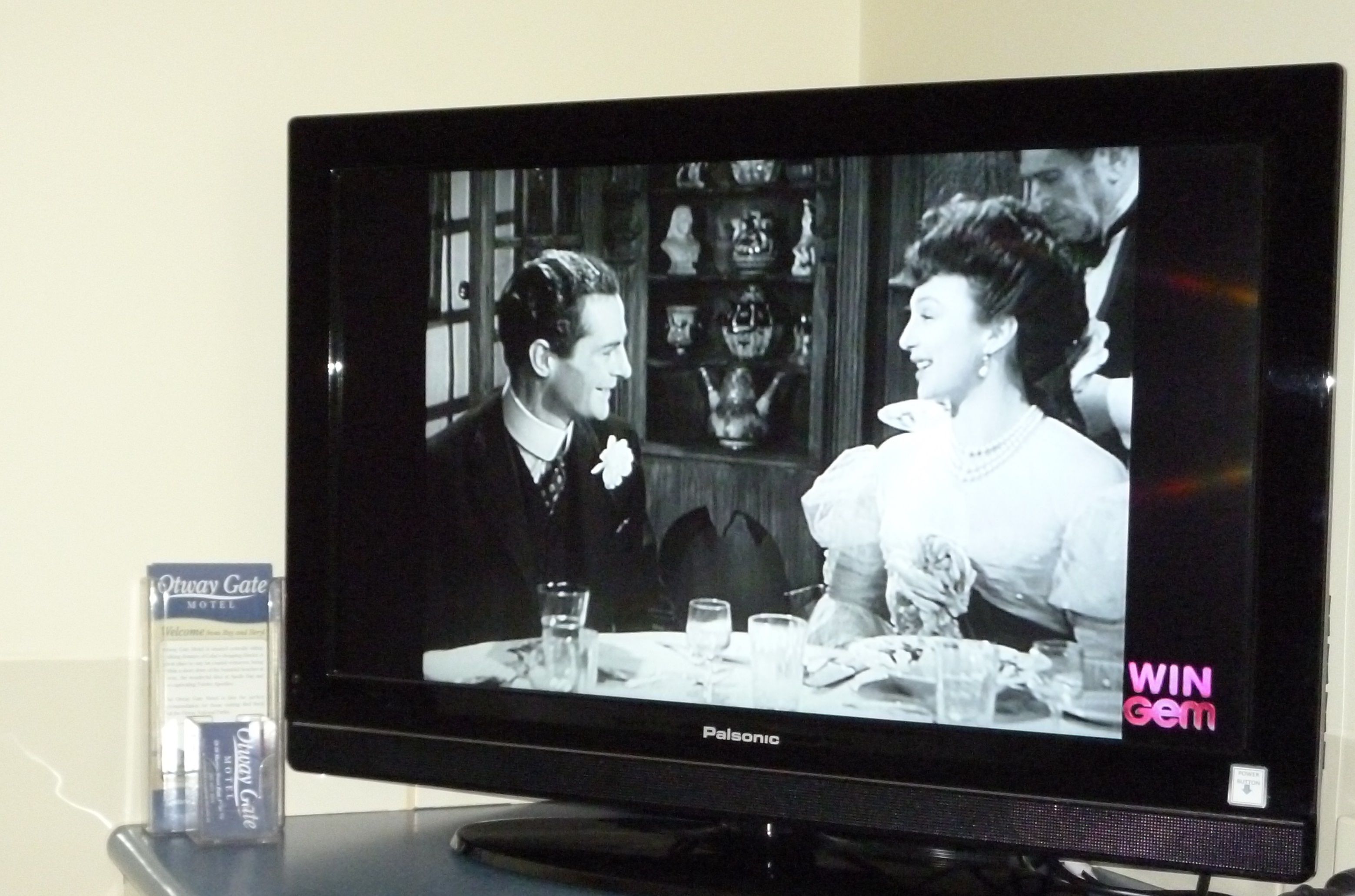 A television displaying a scene with two people at a table, a box labeled "Owyan Gate Motel," and the text "WIN Gem" on the TV screen.