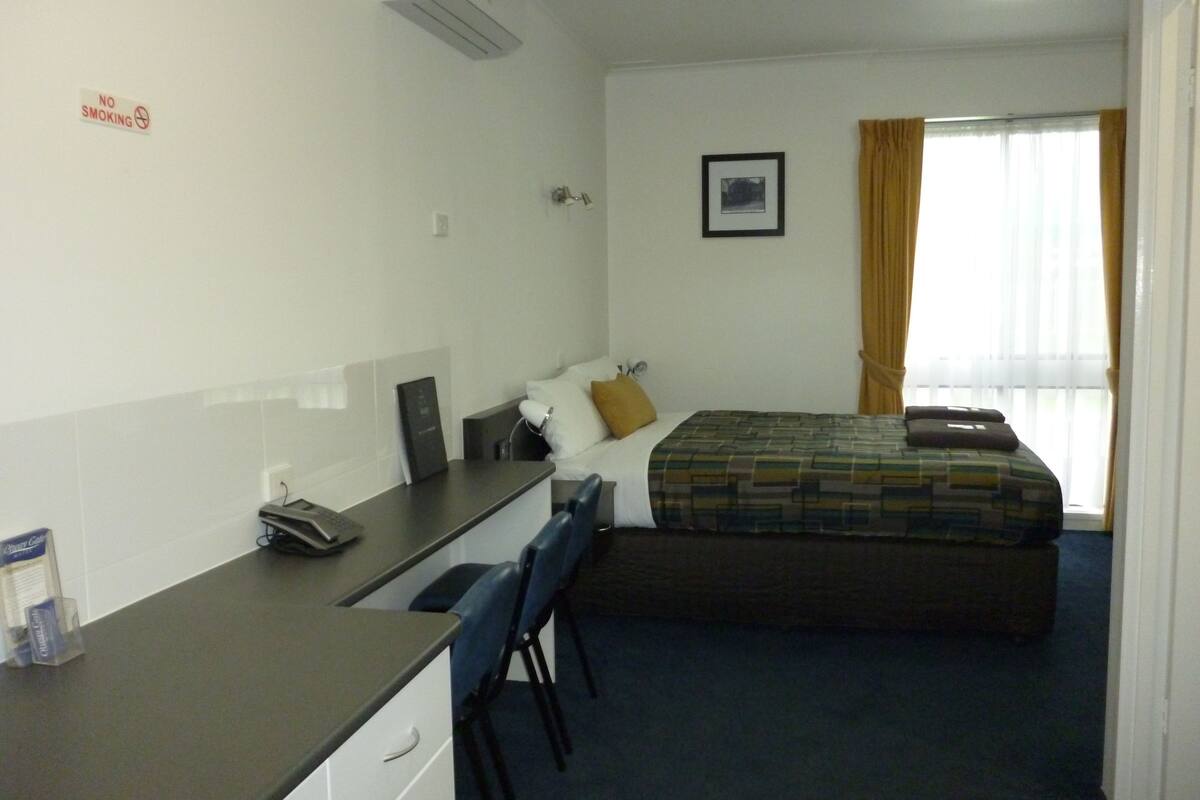 Une chambre d’hôtel comprenant un lit, un bureau avec une chaise, une table de chevet, un tableau au mur et une fenêtre avec des rideaux.