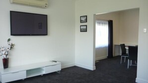 Una habitación con alfombra oscura, una consola blanca con un florero y un televisor de pantalla plana, además de dos cuadros en la pared.