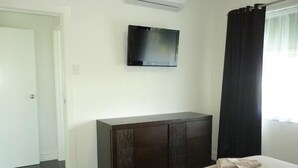 Habitación de hotel con cama, cómoda de madera oscura, televisor de pantalla plana montado en la pared y ventana con cortinas negras.
