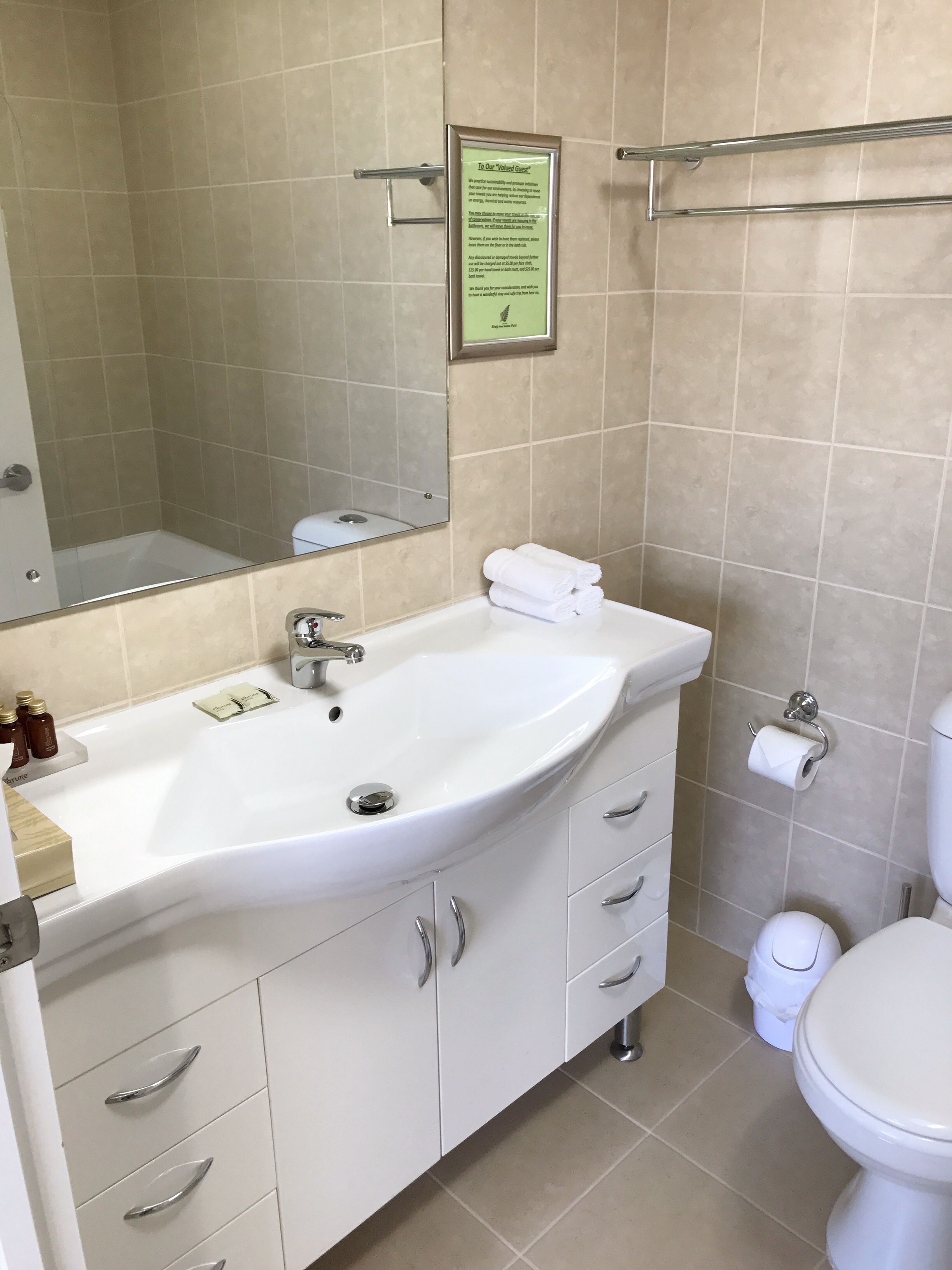A bathroom with a white sink, mirror, and toilet. There is a framed sign on the wall and a towel rack above the sink.