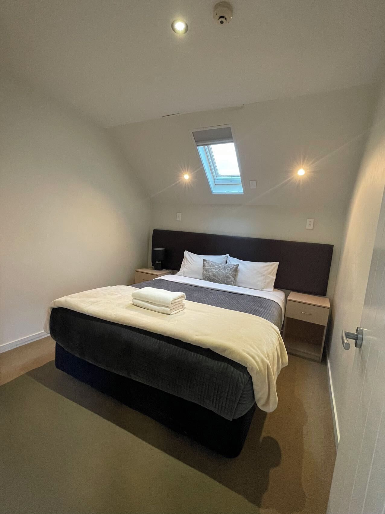 Un dormitorio con tragaluz, una cama con cabecero oscuro, una mesita de noche y una mesilla.