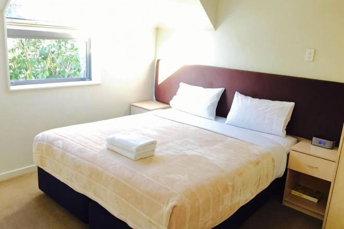 Un lit bien fait, avec une couette blanche et des oreillers, une table de chevet avec une lampe, et une fenêtre donnant sur un paysage verdoyant.