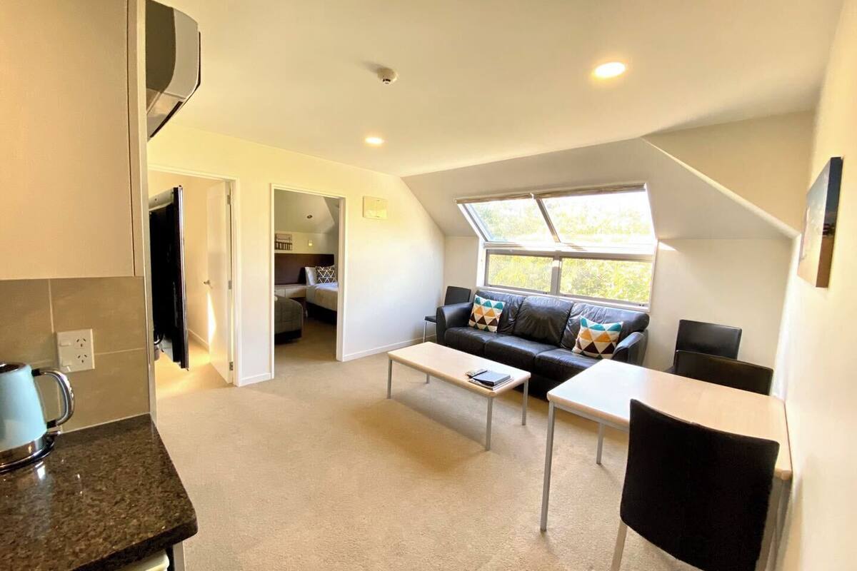 Un espace de vie compact comprenant un coin cuisine, un ensemble de canapés, une table à manger et une grande fenêtre laissant entrer la lumière naturelle.