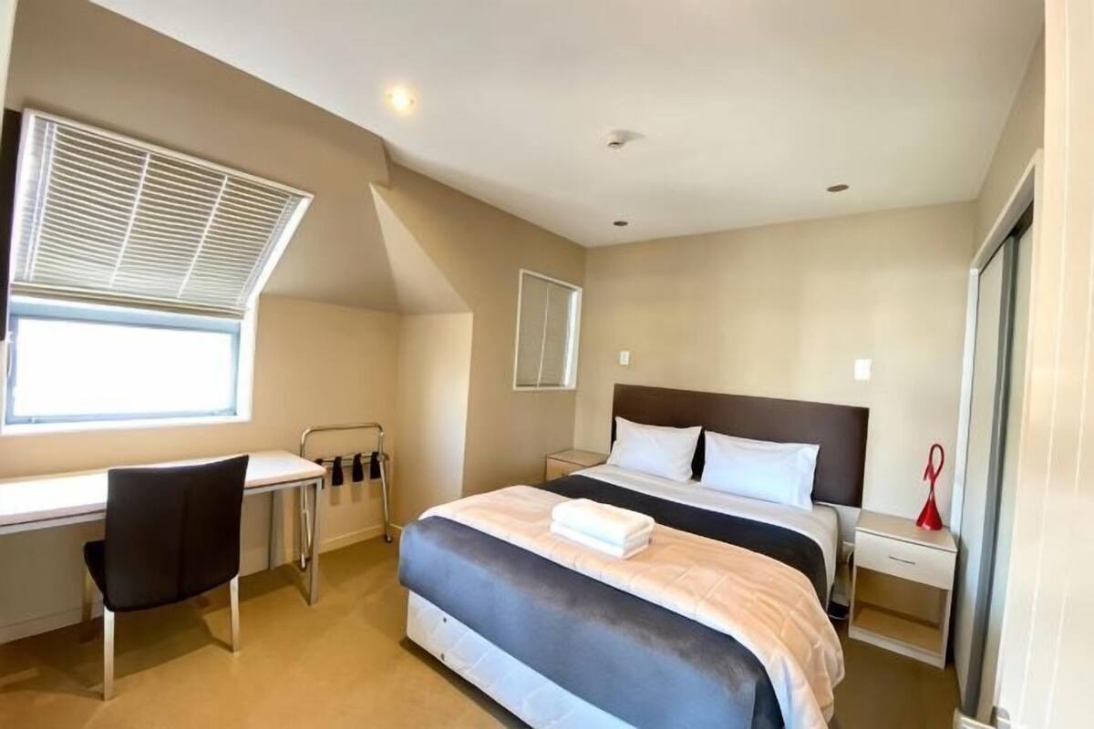 Une chambre à coucher avec un lit, un bureau, une chaise et une fenêtre avec des stores.