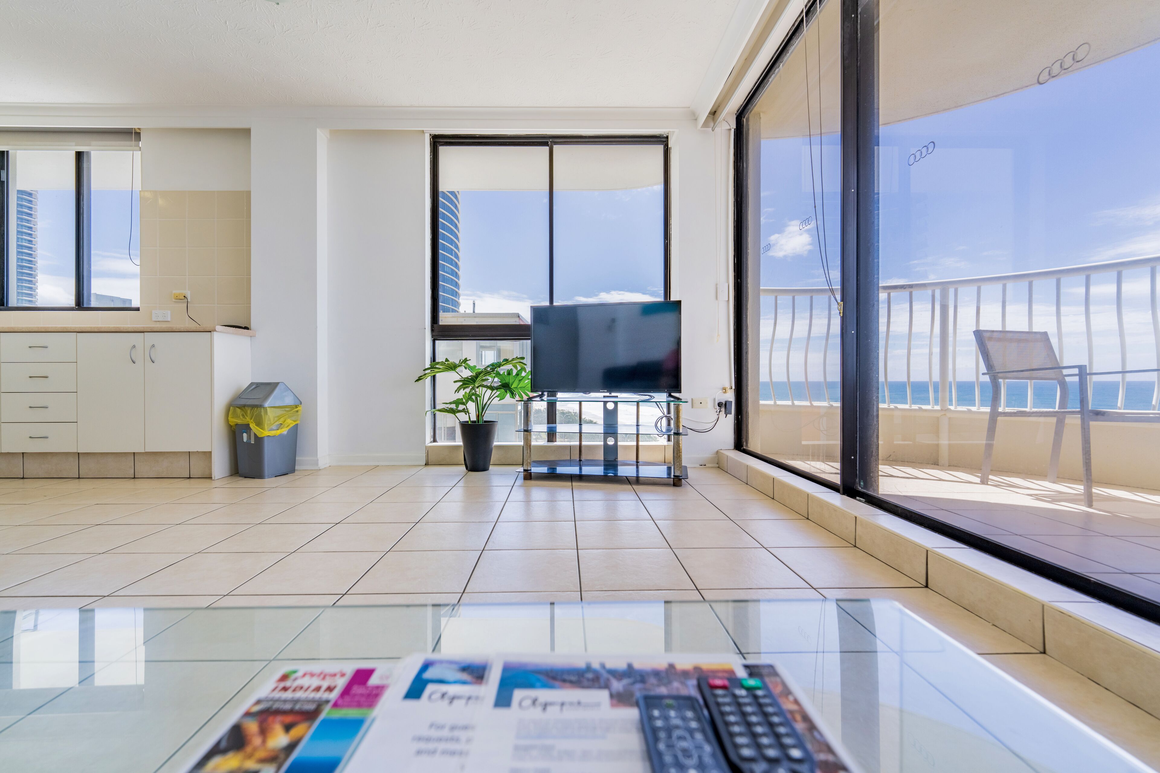 A modern hotel room with a balcony, a TV, a plant, and a glass table with a remote control.