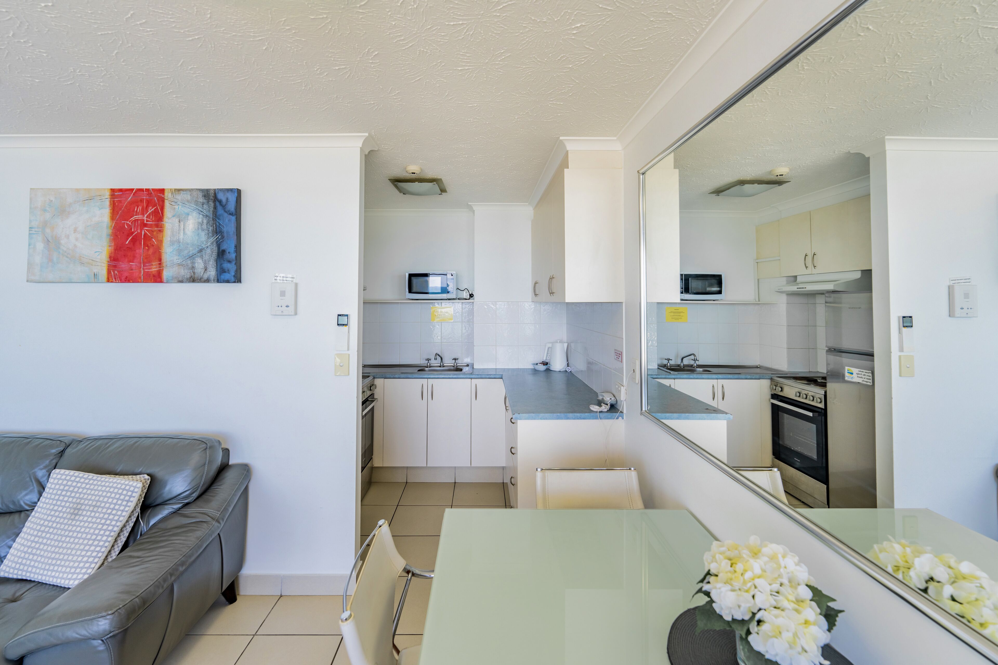 A compact living space with a kitchenette, a grey sofa, a glass dining table, and a decorative piece on the wall.