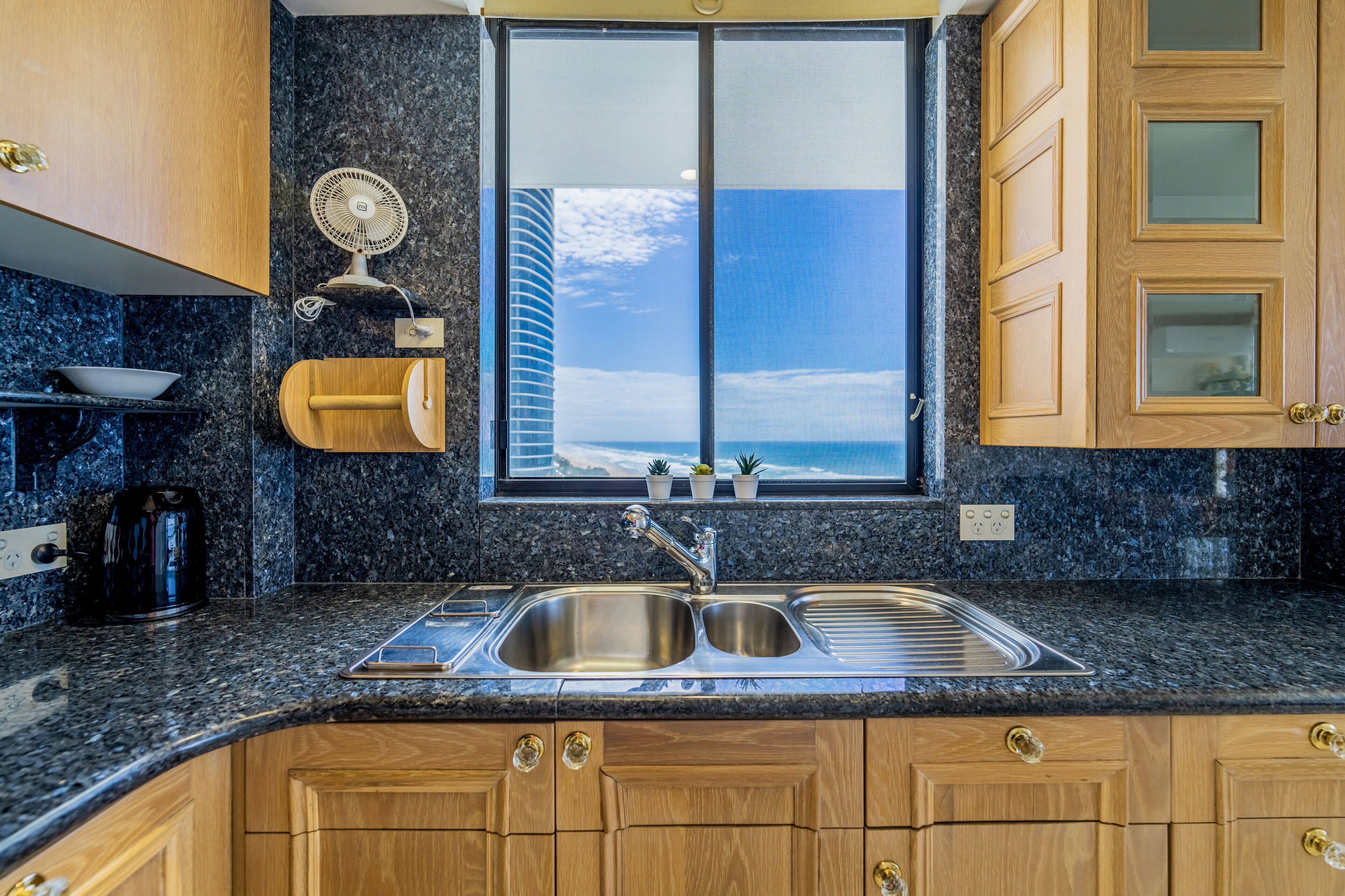 A kitchen with granite countertops, a double sink, wooden cabinets, a built-in fan, and a window with a view of a building and sky.