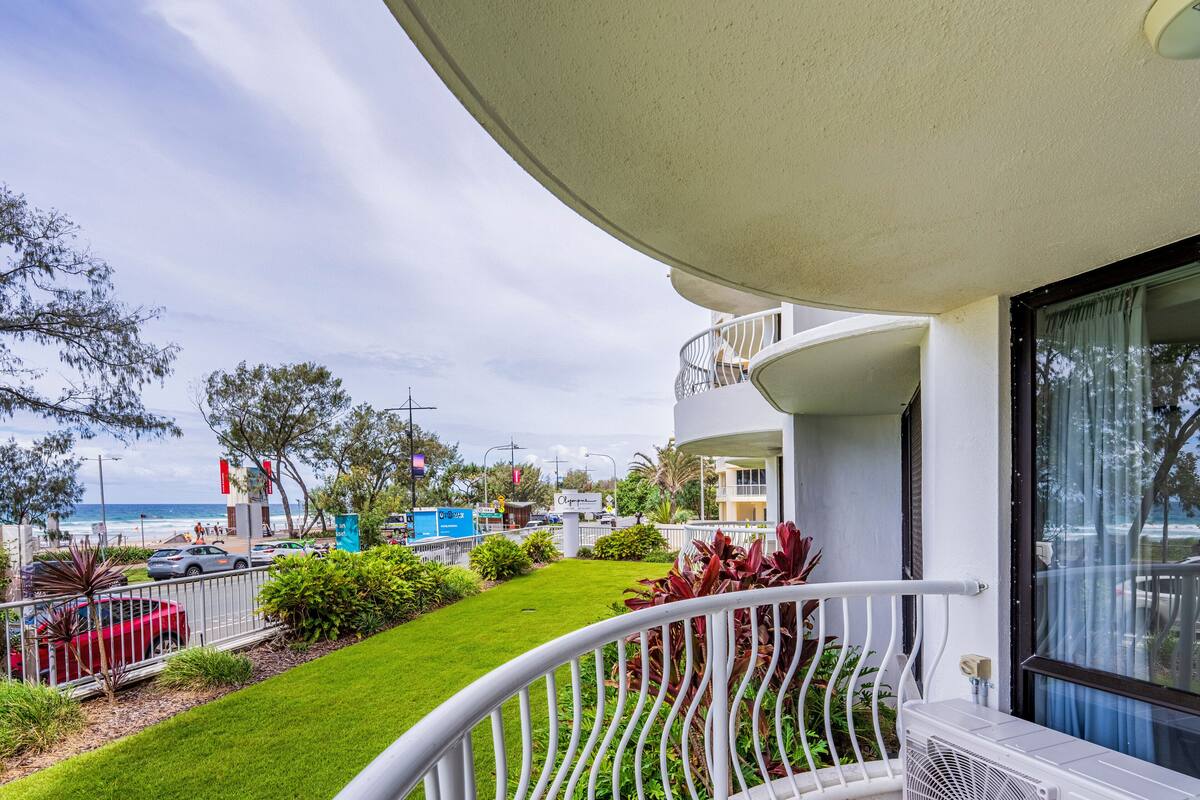 A balcony with a view of the beach and ocean, a well-maintained lawn, and a white railing.