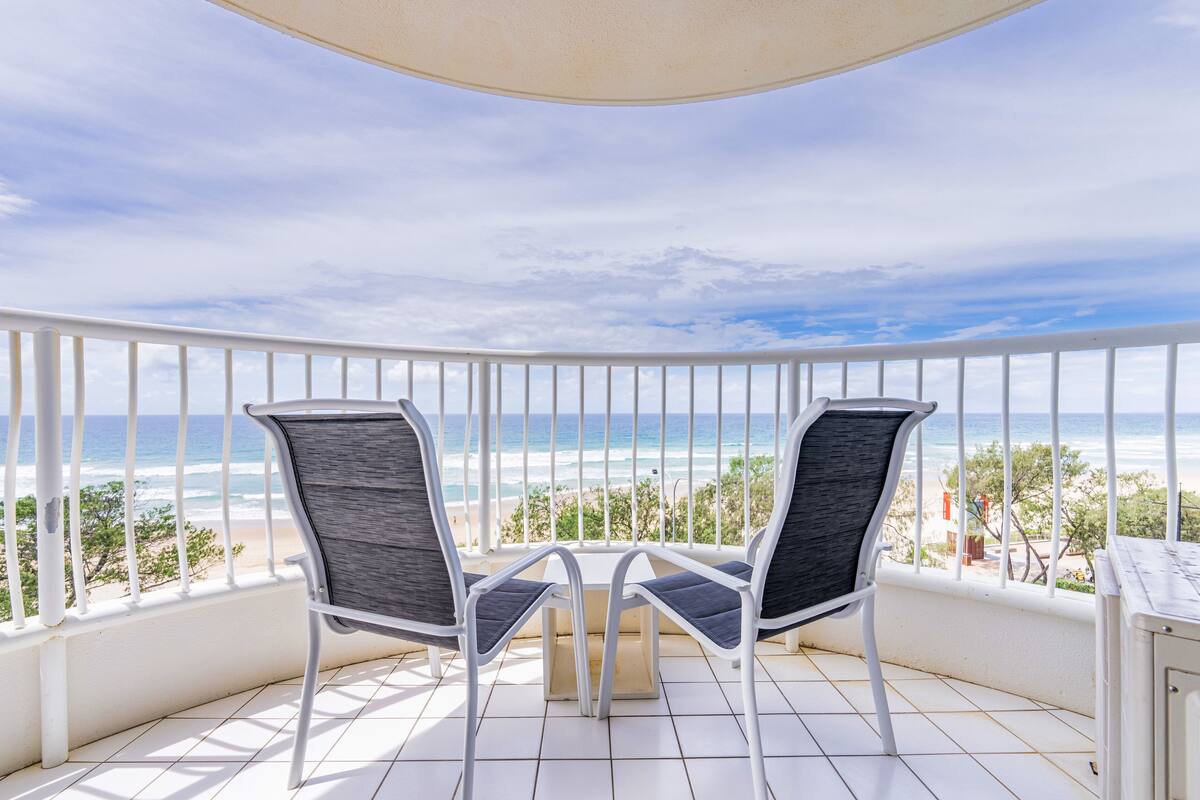 A balcony with two chairs and a table, overlooking a beach with the ocean and sky.