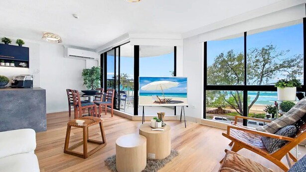 A modern living room with a large window overlooking a beach, a dining area with chairs, and a kitchen area with a counter and plants.