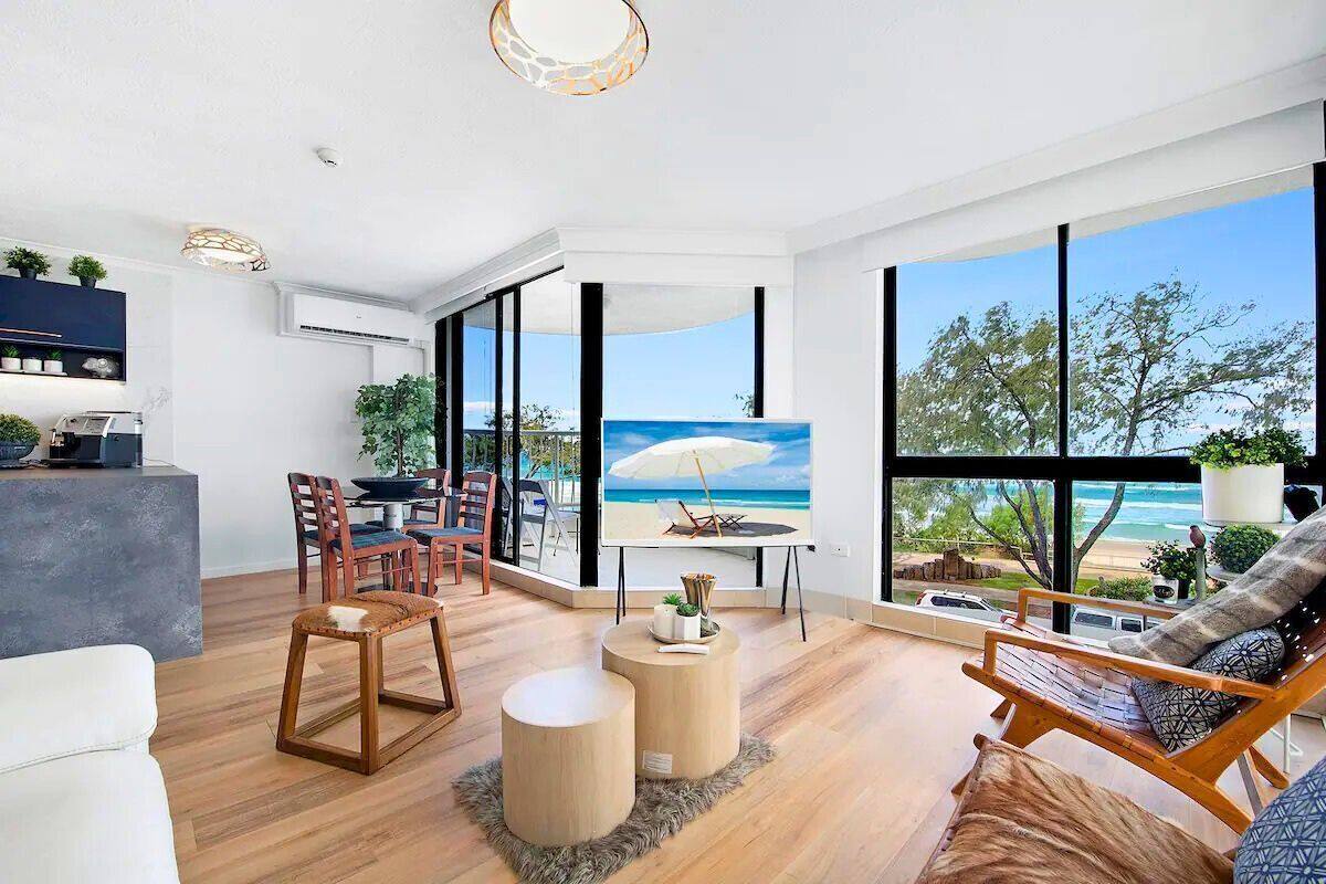 A modern living room with a large window overlooking a beach, a dining area with chairs, and a kitchen area with a counter and plants.