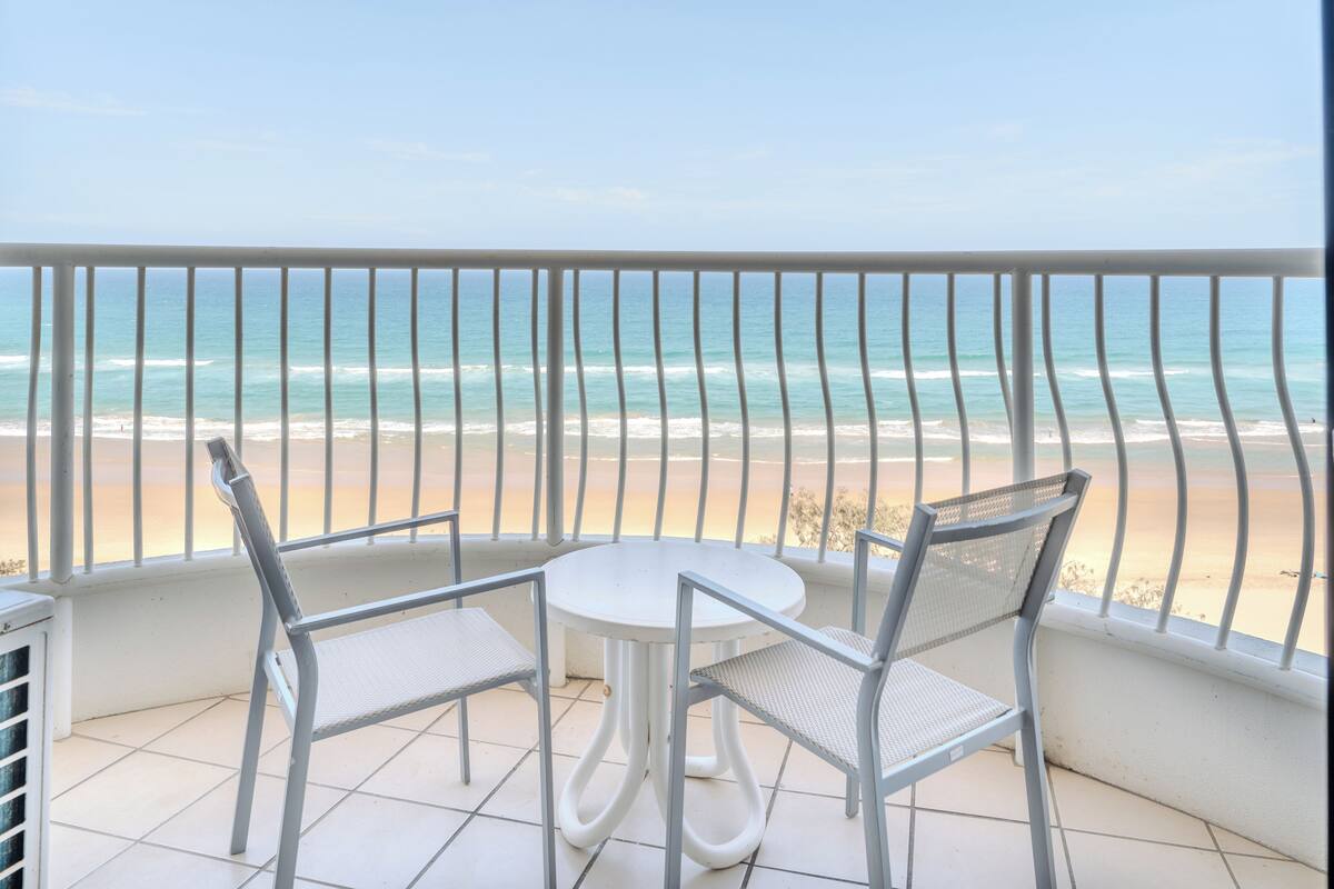 A balcony with a white table and two chairs, overlooking a beach with clear blue water and sandy shore.