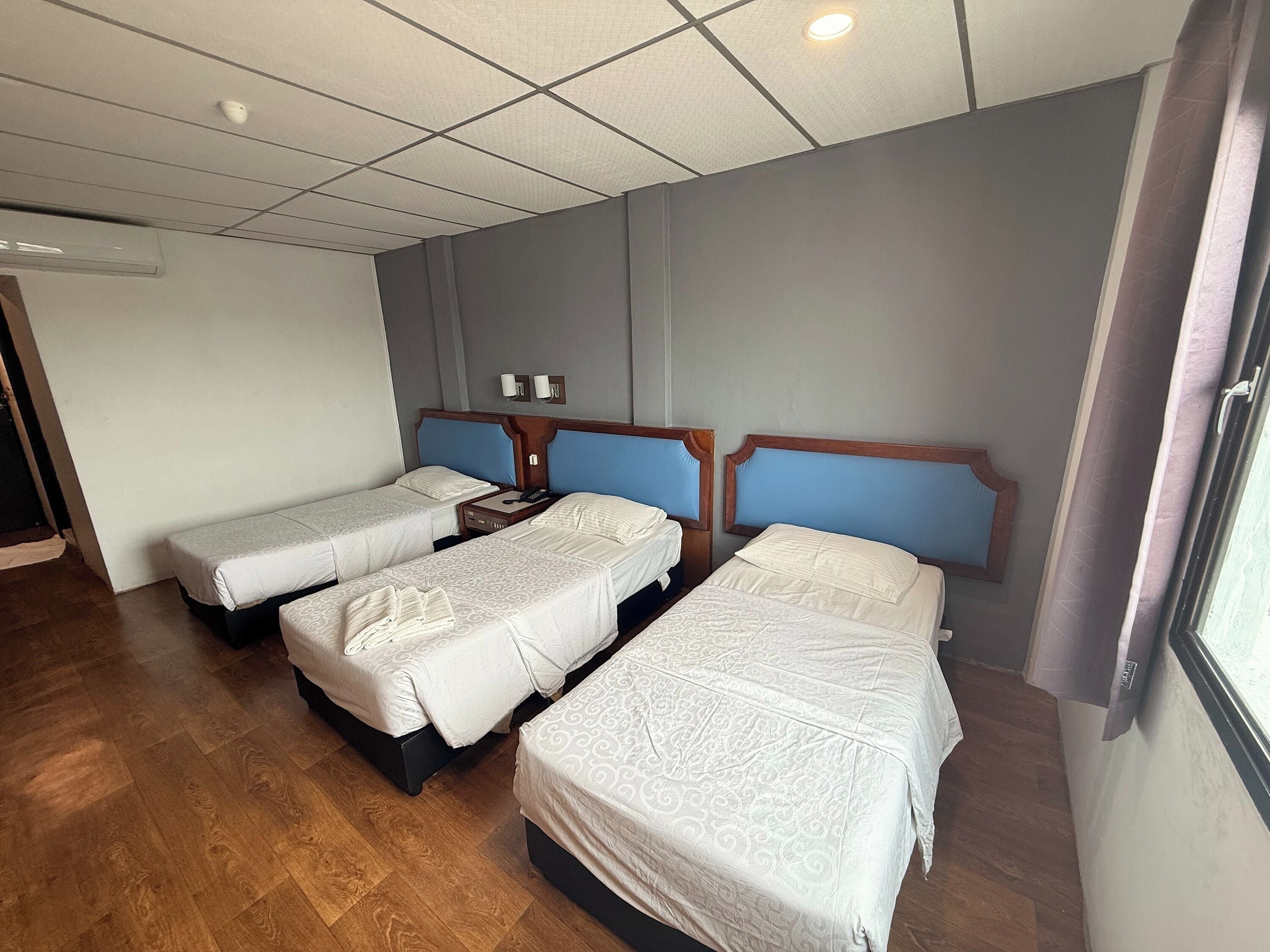 A hotel room with two single beds and one double bed, wooden flooring, a ceiling with recessed lighting, and a window with curtains.