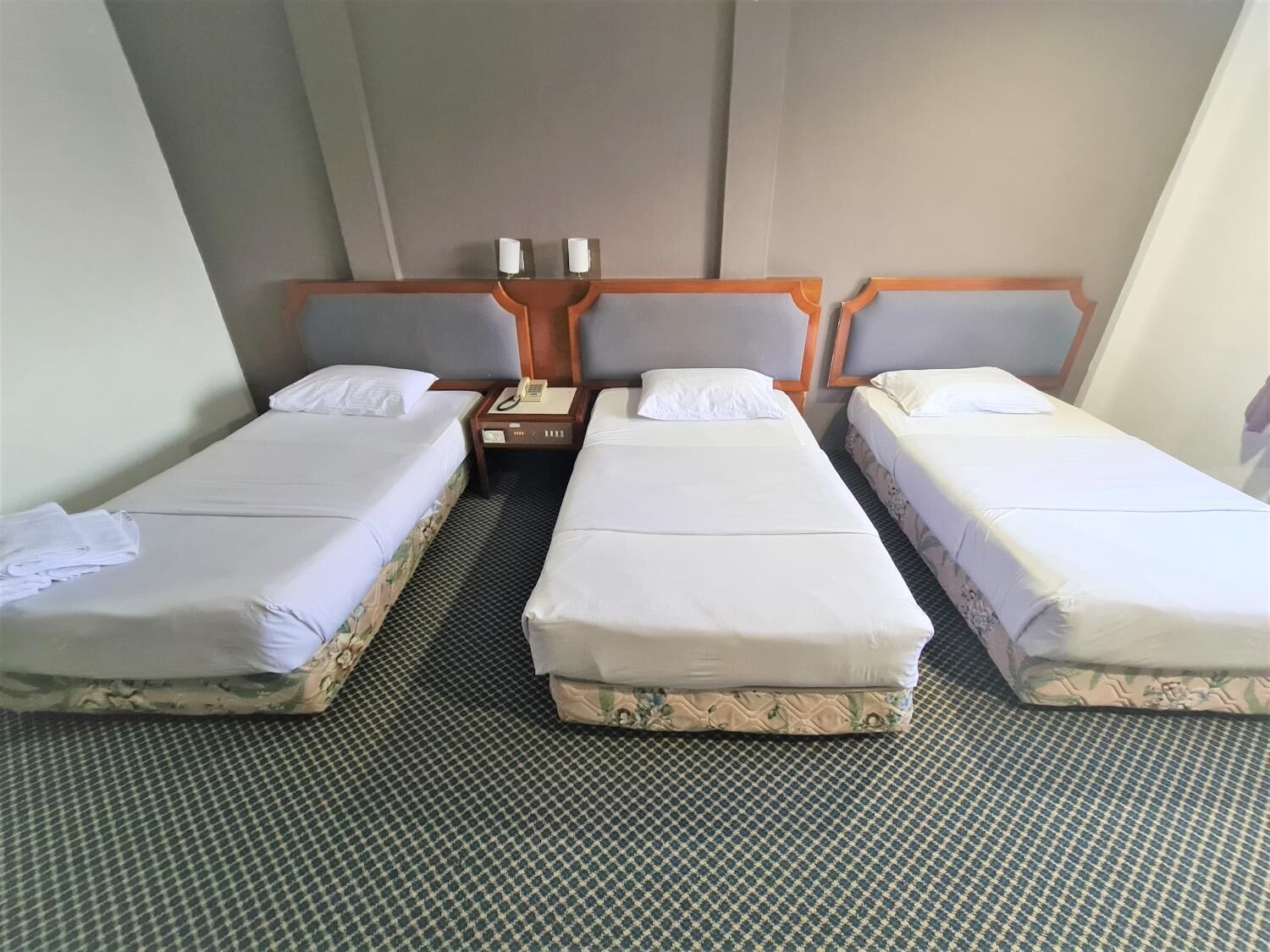 Habitación de hotel con tres camas individuales, una mesita de noche de madera con un teléfono y una alfombra estampada.