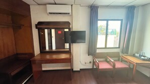 A hotel room with a wooden desk, a chair, a small table, an air conditioning unit, a window with curtains, and a view of the outside.