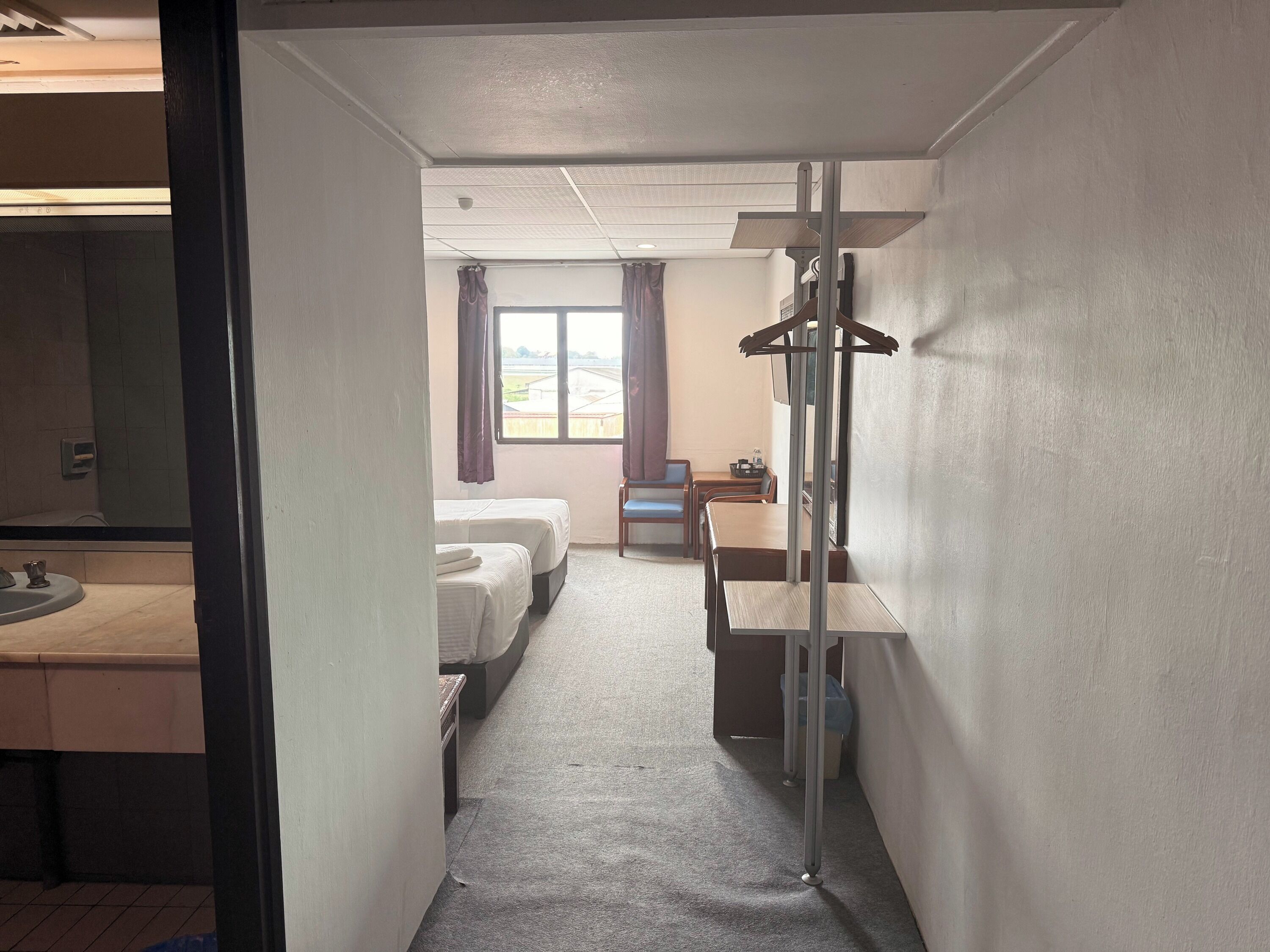 Ein Hotelzimmer mit zwei Betten, einem Schreibtisch, einem Stuhl, einem kleinen Tisch und einem Fenster mit Vorhängen.