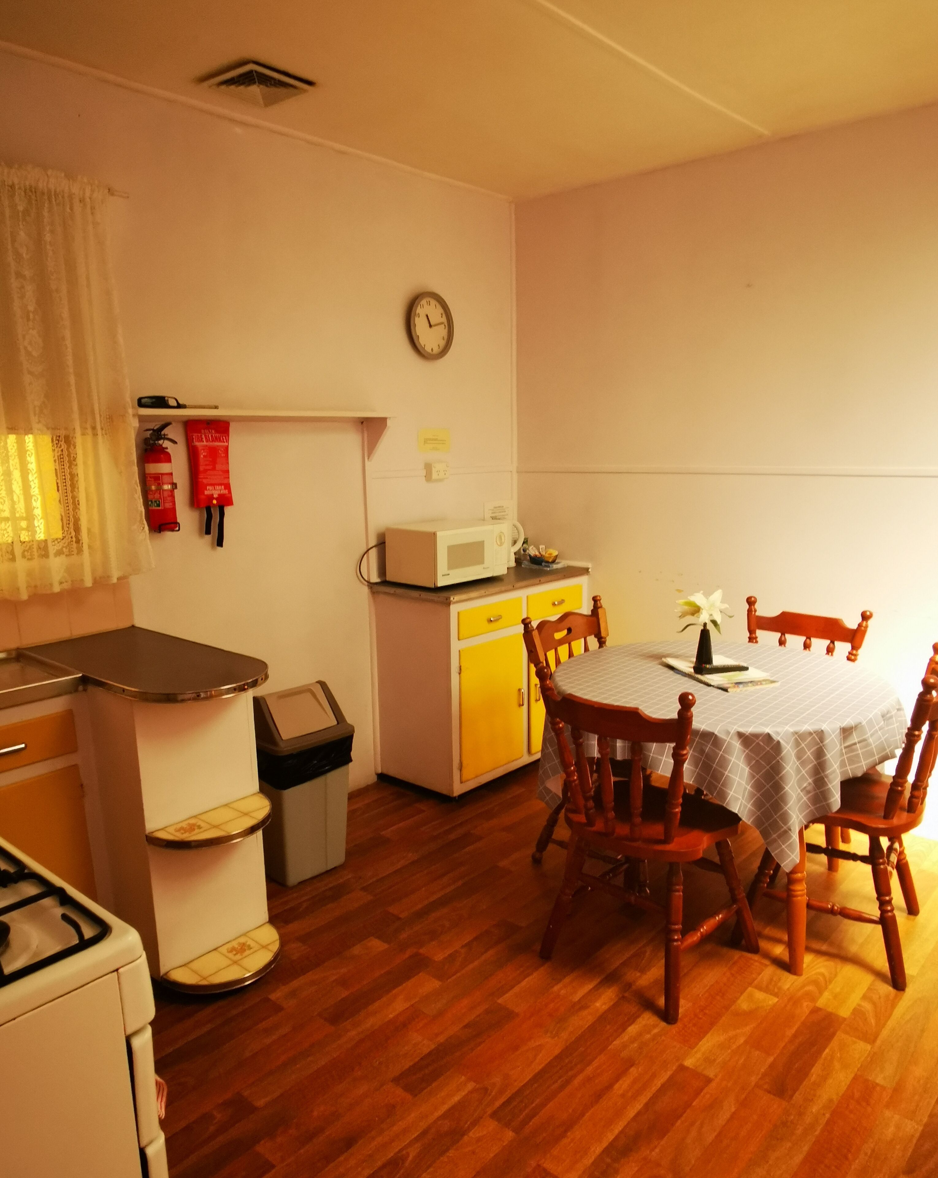 A compact kitchen with a microwave, a small refrigerator, and a dining table with chairs. There is a clock on the wall and a fire extinguisher on the wall.