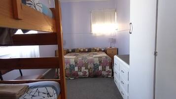 A dormitory room with bunk beds, a window with curtains, a bedside table, and a wardrobe.