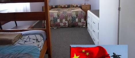 A dormitory room with bunk beds, a window with curtains, a bedside table, and a wardrobe.
