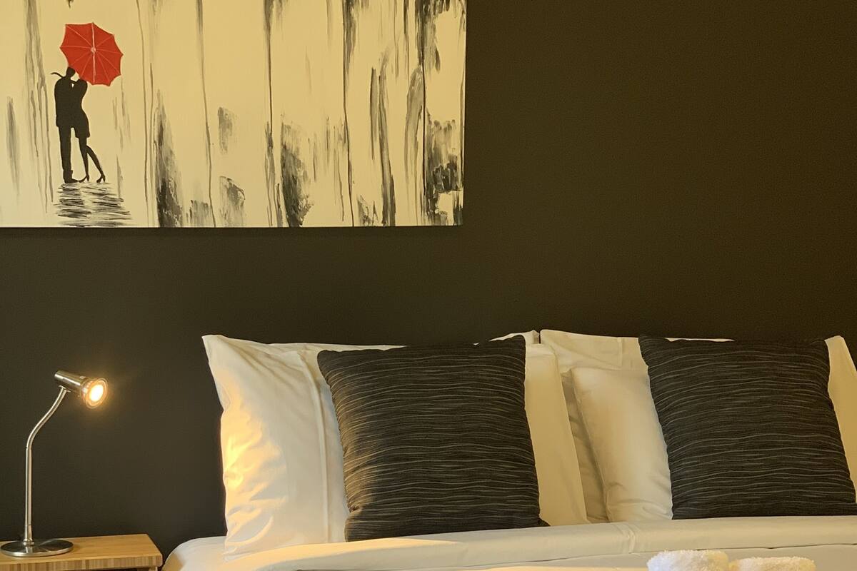 Ein ordentlich bezogenes Bett mit weißen Laken, einer roten Tagesdecke und einem Nachttisch mit Lampe.